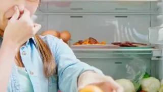 Someone in front of a fridge holding her nose with one hand and a rotten orange in the other hand.