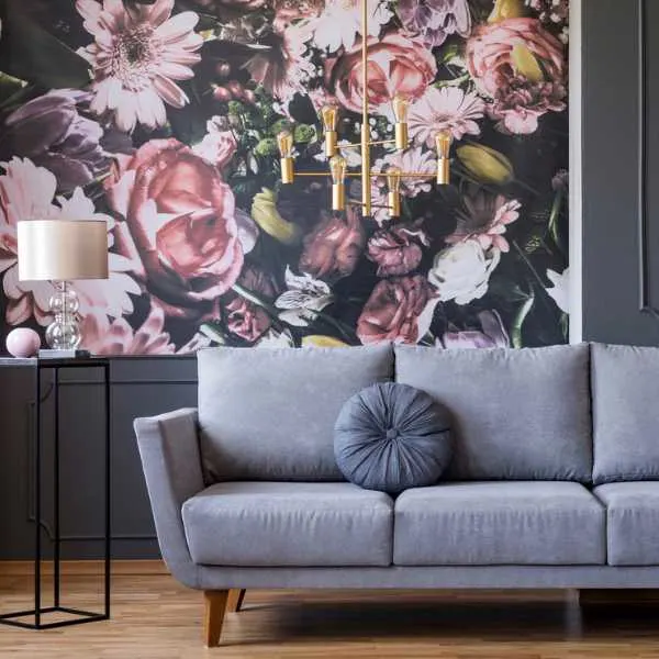 Gray couch and table lamp in front of floral wallpaper.