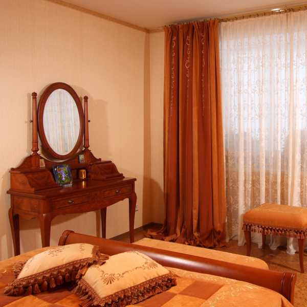 Bedroom with brown dresser, cream wall and burnt orange drape.